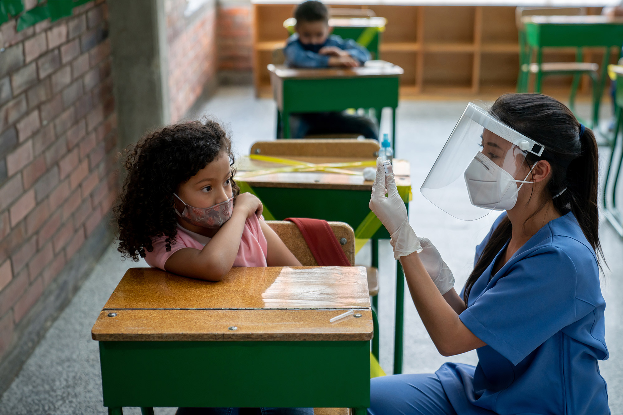 A woman wearing blue scrubs, a mask, and a face shield draws medication from a vile. A child with curly brown hair and wearing a face mask sits at a desk in a class room and leans away from the woman.