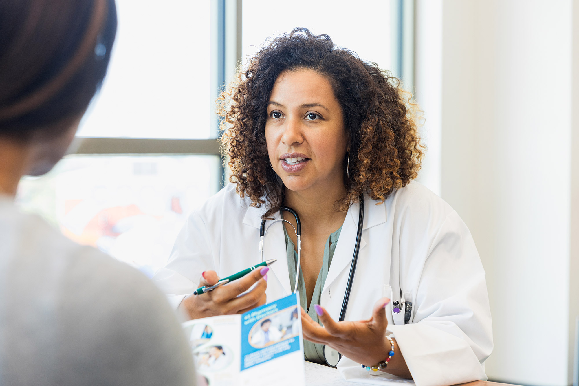 A black woman with curly brown hair, wearing a white coat and a stethoscope around her neck, sits and gestures with her hands as she speaks to a woman seated across from her.