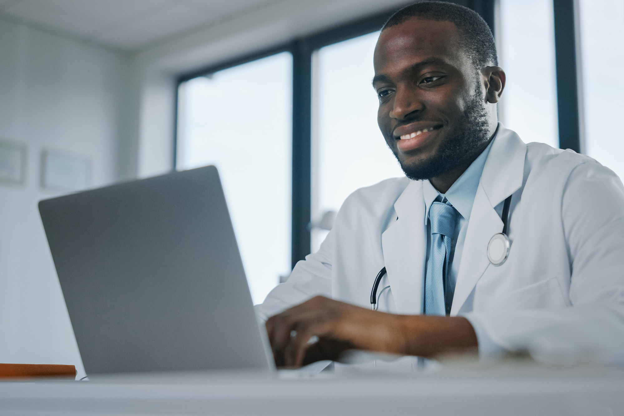 A smiling black man wearing a white coat and a stethoscope around his neck sitting at a desk typing on a laptop.