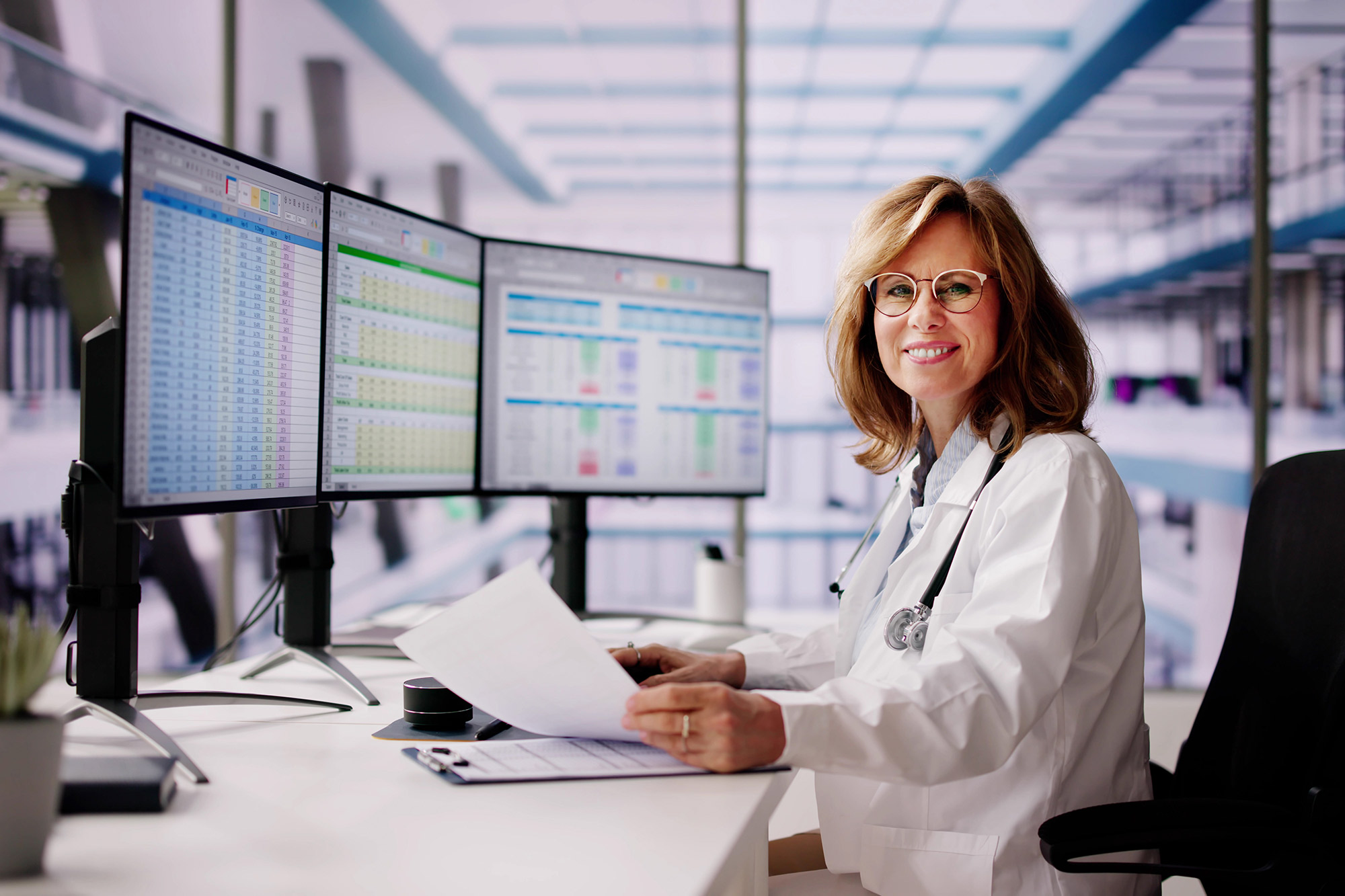 A middle aged woman with blonde hair wearing a white coat and a stethoscope around her neck sits at a desk with three large computer monitors.