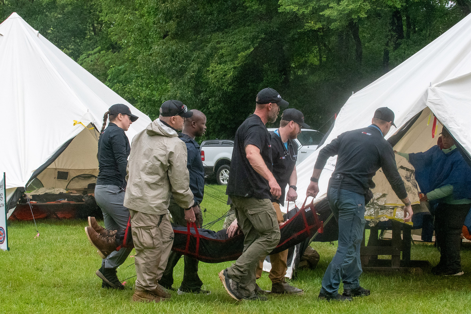 A diverse group of men and and one woman wearing black baseball caps carry a man on a litter between two large white tents.