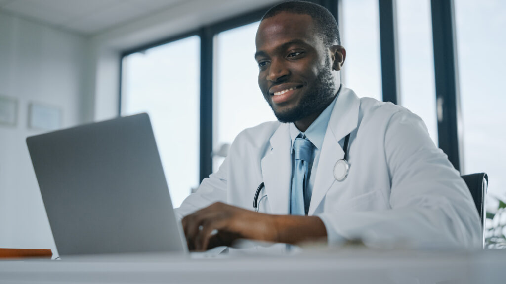 Happy African American Medical Doctor is Working on a Laptop Computer in a Health Clinic.