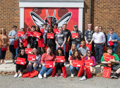 A large group of students pose for a photo holding up red envelopes that say "you're in!"