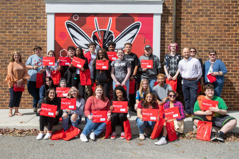 A large group of students pose for a photo holding up red envelopes that say "you're in!"
