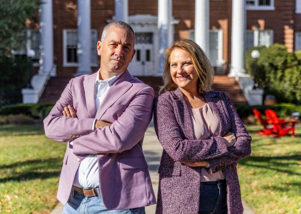 A man and a woman pose for a photo. Both are wearing light purple sports coats and both are crossing their arms over their chests.