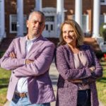 A man and a woman pose for a photo. Both are wearing light purple sports coats and both are crossing their arms over their chests.