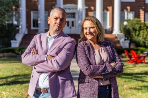 A man and a woman pose for a photo. Both are wearing light purple sports coats and both are crossing their arms over their chests.