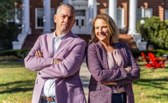 A man and a woman pose for a photo. Both are wearing light purple sports coats and both are crossing their arms over their chests.
