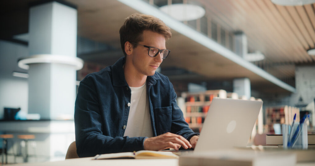 Concentrated Young Caucasian Male Student Working on His Laptop in a Modern Library. Man Engaged in Academic Research With Books Around, Focused on Screen.