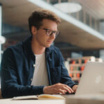 Concentrated Young Caucasian Male Student Working on His Laptop in a Modern Library. Man Engaged in Academic Research With Books Around, Focused on Screen.