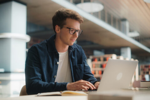 Concentrated Young Caucasian Male Student Working on His Laptop in a Modern Library. Man Engaged in Academic Research With Books Around, Focused on Screen.