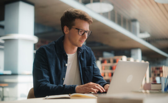 Concentrated Young Caucasian Male Student Working on His Laptop in a Modern Library. Man Engaged in Academic Research With Books Around, Focused on Screen.