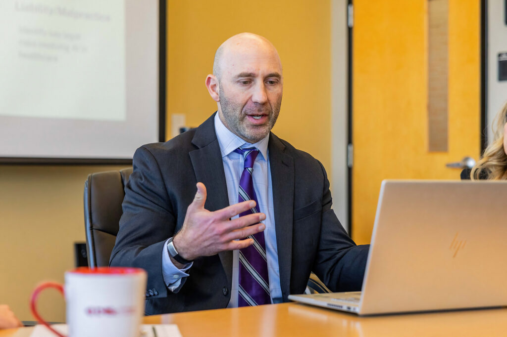 A man in a suit sits gestures as he sits in front of a laptop.