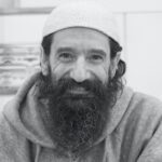 A man with a long beard, wearing a religious head covering and a gray hoodie, smiles at the camera.