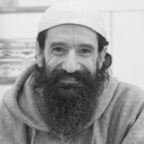 A man with a long beard, wearing a religious head covering and a gray hoodie, smiles at the camera.
