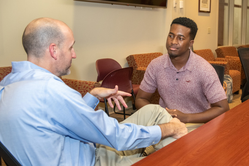 Two people sit and talk in an office lounge area, with one gesturing while the other listens attentively.
