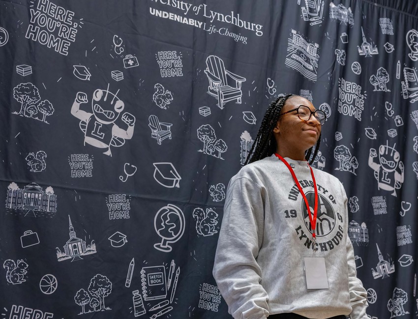 A student wearing a University of Lynchburg sweatshirt and red lanyard stands in front of a branded backdrop covered with campus-themed illustrations and “Here, you’re home” graphics.