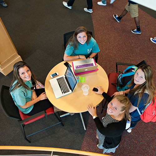 Four students gathered around a round table with laptops, looking up in a campus study space.