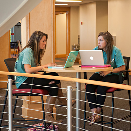 Two people sit at a small table in a campus café area, talking with laptops open.