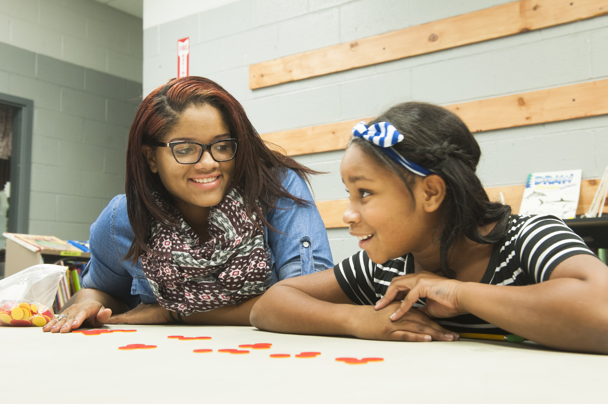 Young woman smiles at a child across a table during a learning activity, with colorful counters spread out in front of them.
