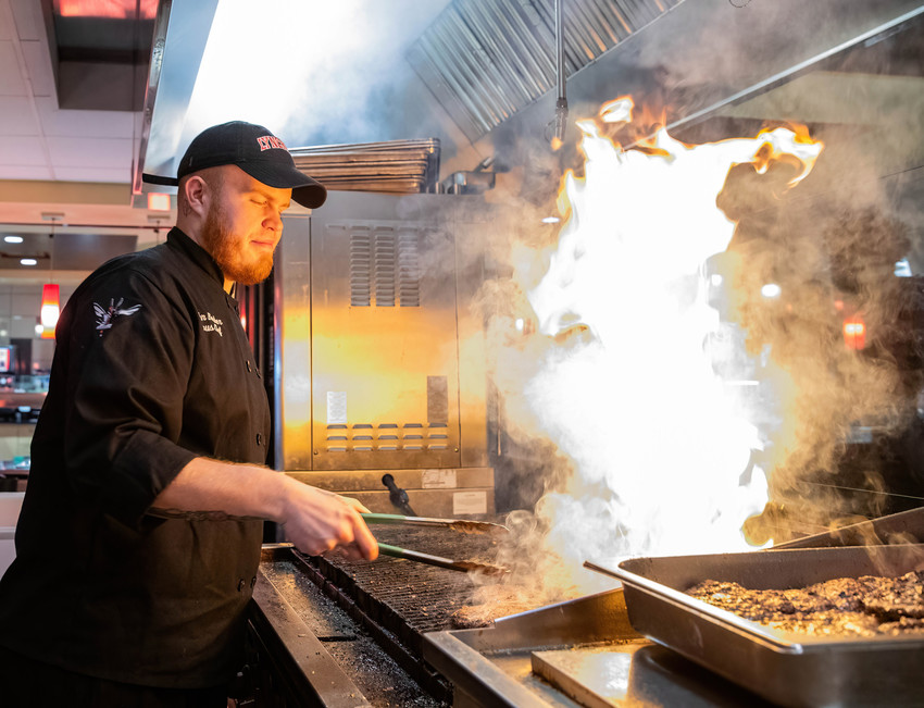 Chef cooks at a campus grill as flames rise from the stovetop in a commercial kitchen.