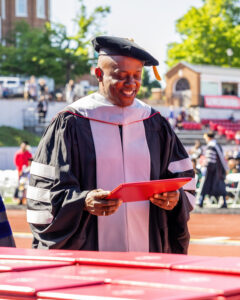 A black man wearing silver doctoral regalia smiles as he holds his diploma. 