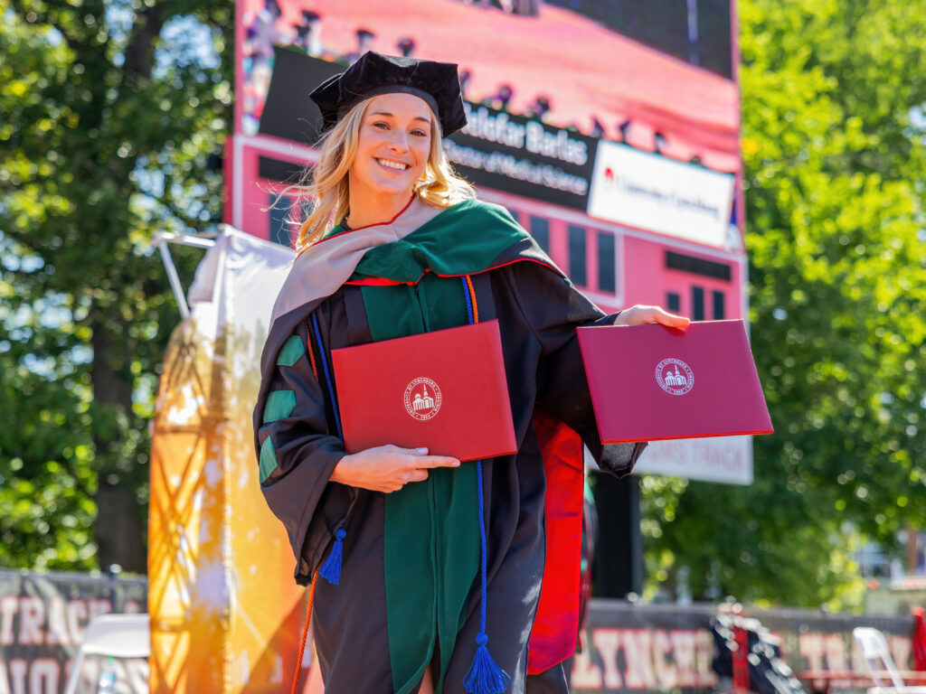 A woman wearing doctoral regalia poses with two diplomas. 