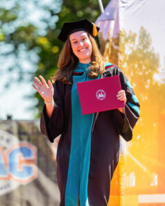 A white woman wearing doctoral regalia waves to the camera and holds up her diploma. 