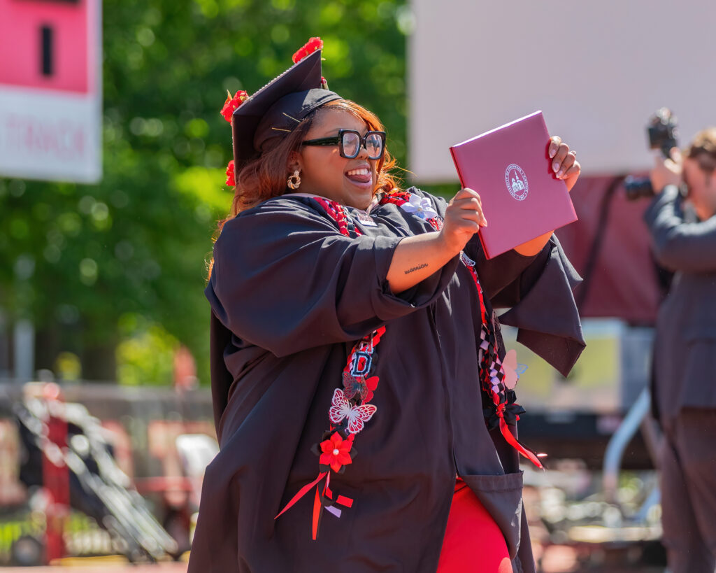 A black woman wearing academic regalia holds up her diploma while sticking out her tongue. 