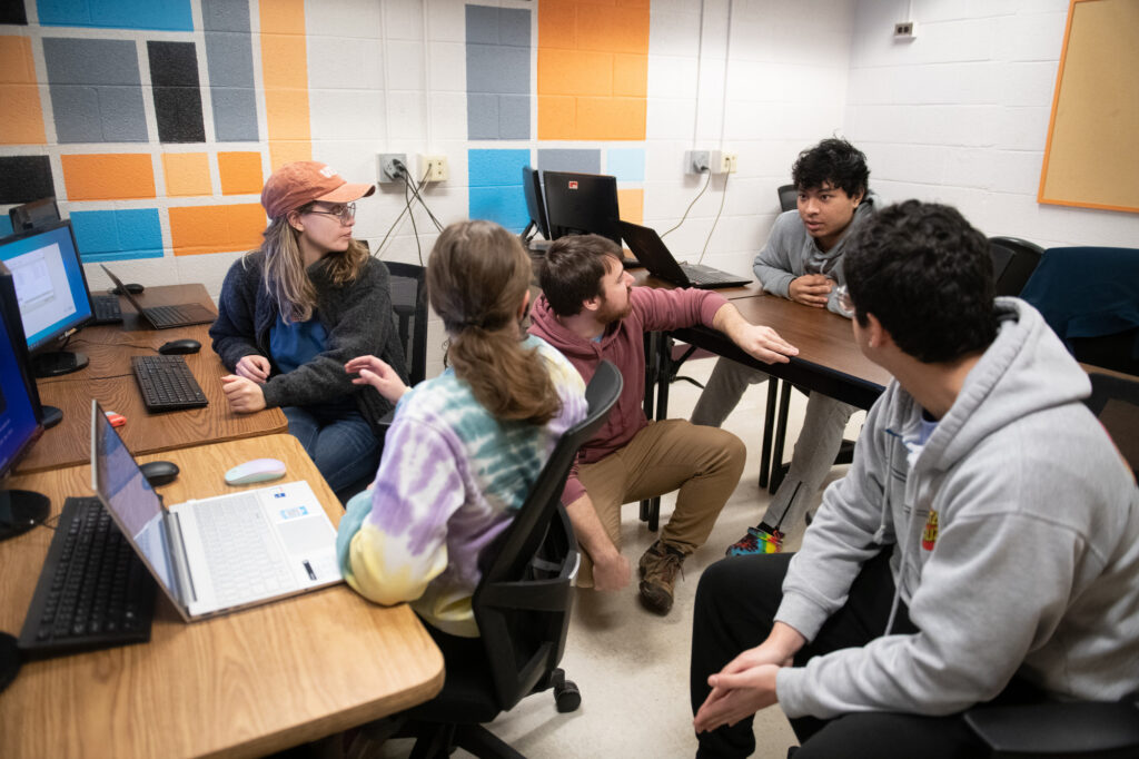 Students collaborate in a computer lab during a cybersecurity minor class, discussing ideas around tables with desktop computers and laptops.