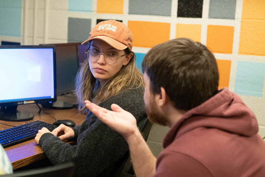 Students in a cybersecurity minor lab discuss work at computer stations, with one student at a desktop computer listening as another gestures during conversation.