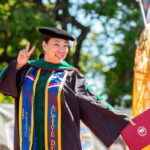 An asian woman wearing green doctoral regalia hold up a peace sign.
