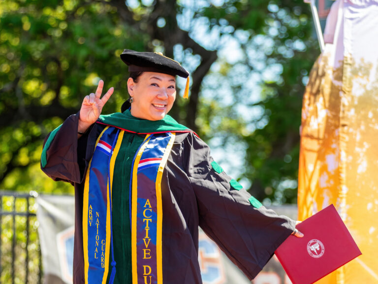 An asian woman wearing green doctoral regalia hold up a peace sign.