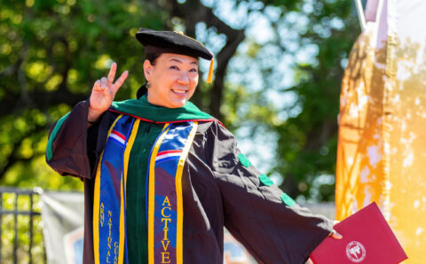 An asian woman wearing green doctoral regalia hold up a peace sign.