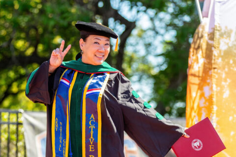 An asian woman wearing green doctoral regalia hold up a peace sign.