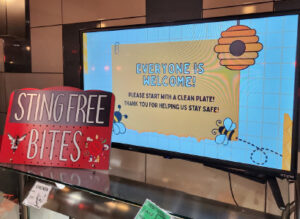 A food service display shows a colorful “Sting Free Bites” sign beside a digital screen that reads “Everyone is welcome! Please start with a clean plate! Thank you for helping us stay safe!” with bee and hive graphics.
