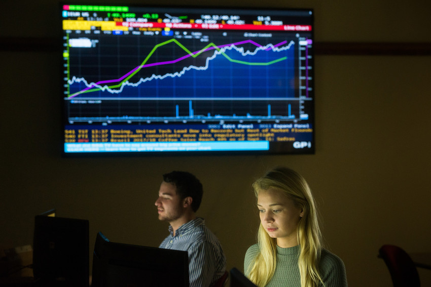 Two students work at computer stations beneath a large financial market chart display, representing finance, investment analysis, and business data monitoring.