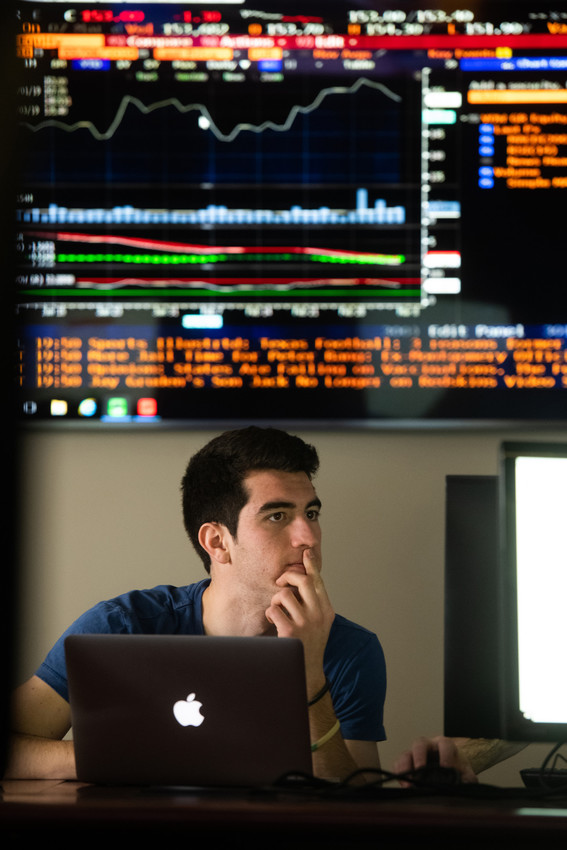 Student studies market data on a laptop beneath a large financial chart display, representing business analytics, finance, and data-driven decision-making.