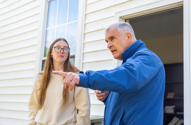Older man points into the distance while speaking with a student outside a light-colored building.