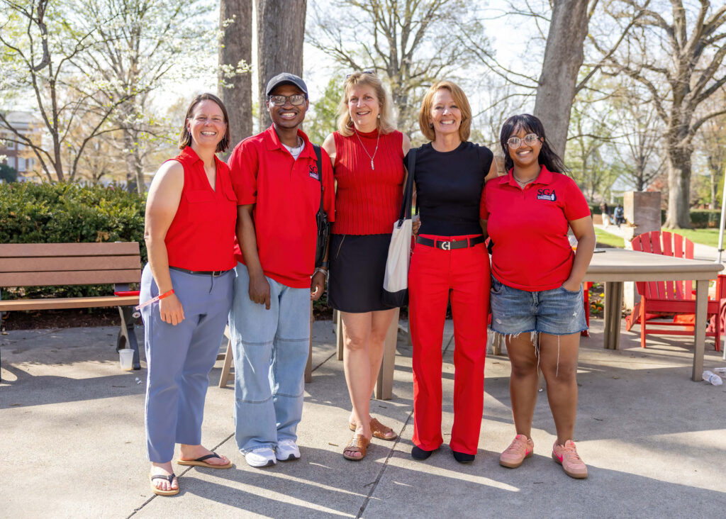 Five smiling students and staff members stand together outdoors on campus, posing for a group photo near benches and red Adirondack chairs.