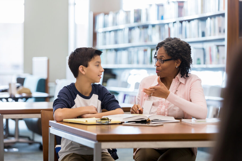 A middle school age boy sits at a table in a school library with his mature female mentor. He listens as she gestures and speaks.