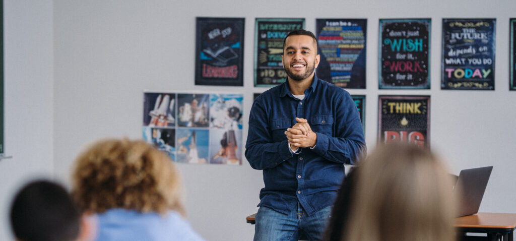 A male teacher of Hispanic ethnicity stands in the front of his middle school classroom, teaching a lesson to a group of concentrated students.
