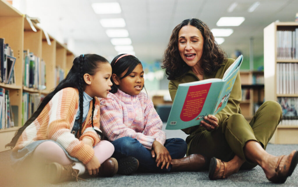 Girl, teacher and book with reading at library for development, learning or study with guide. School kids, storytelling and woman with help on floor, education and language for knowledge at academy
