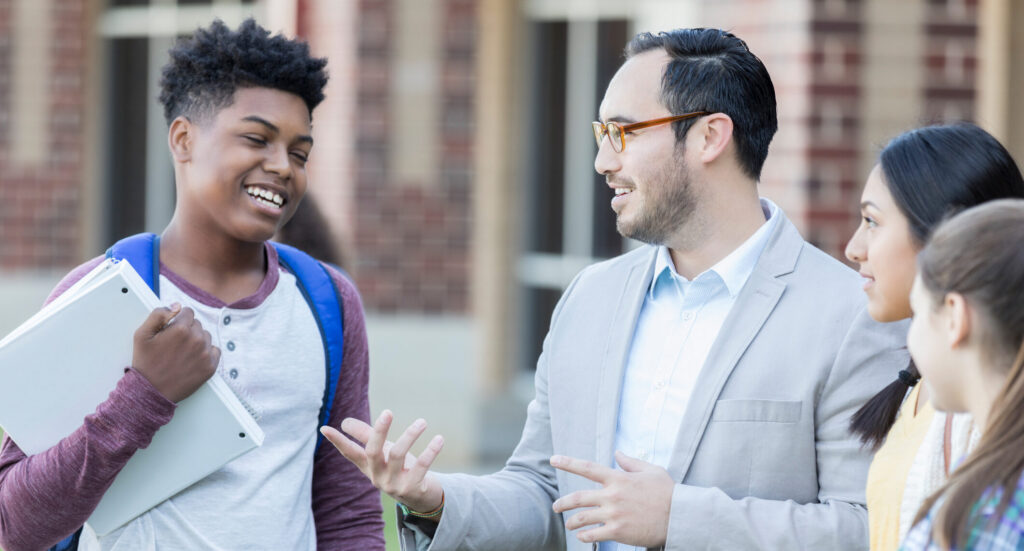 A mid adult high school teacher talks with his hands as he discusses his latest lessons with students between classes. Three students stand with him outside.