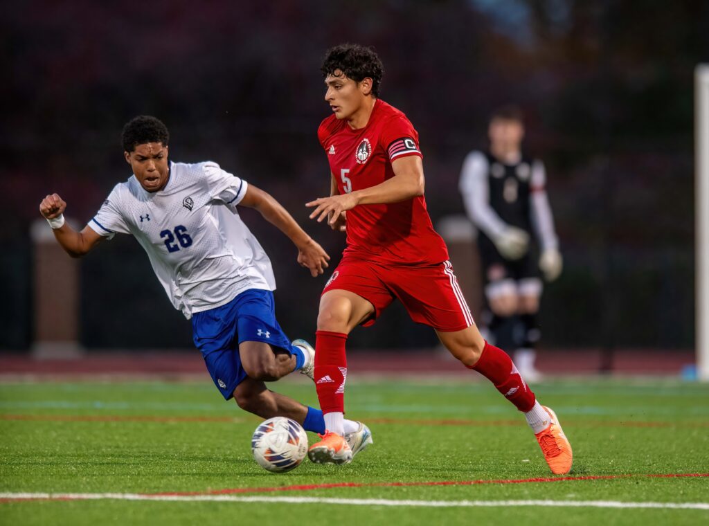 Men's Soccer Player in a red jersey, kicking the ball past an opposing player in a white and blue jersey.
