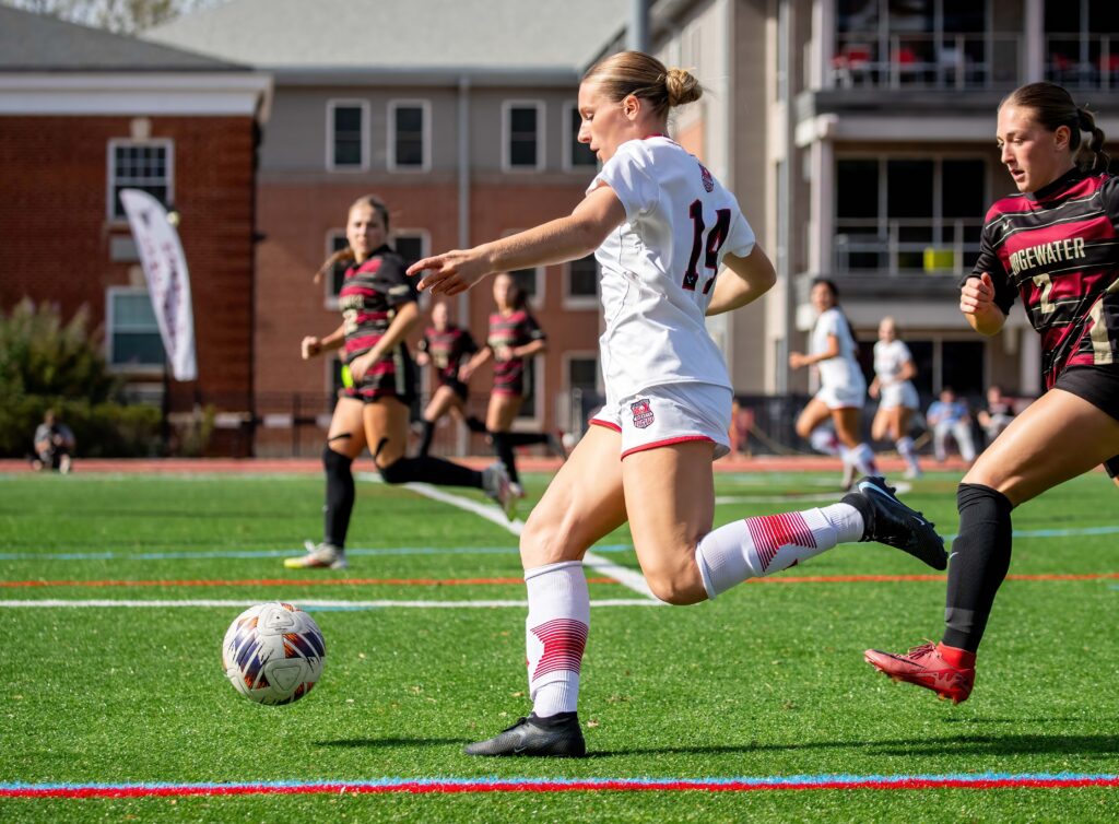 lynchburg women's soccer player in a white jersey chases after the ball and passes past an opposing defender.