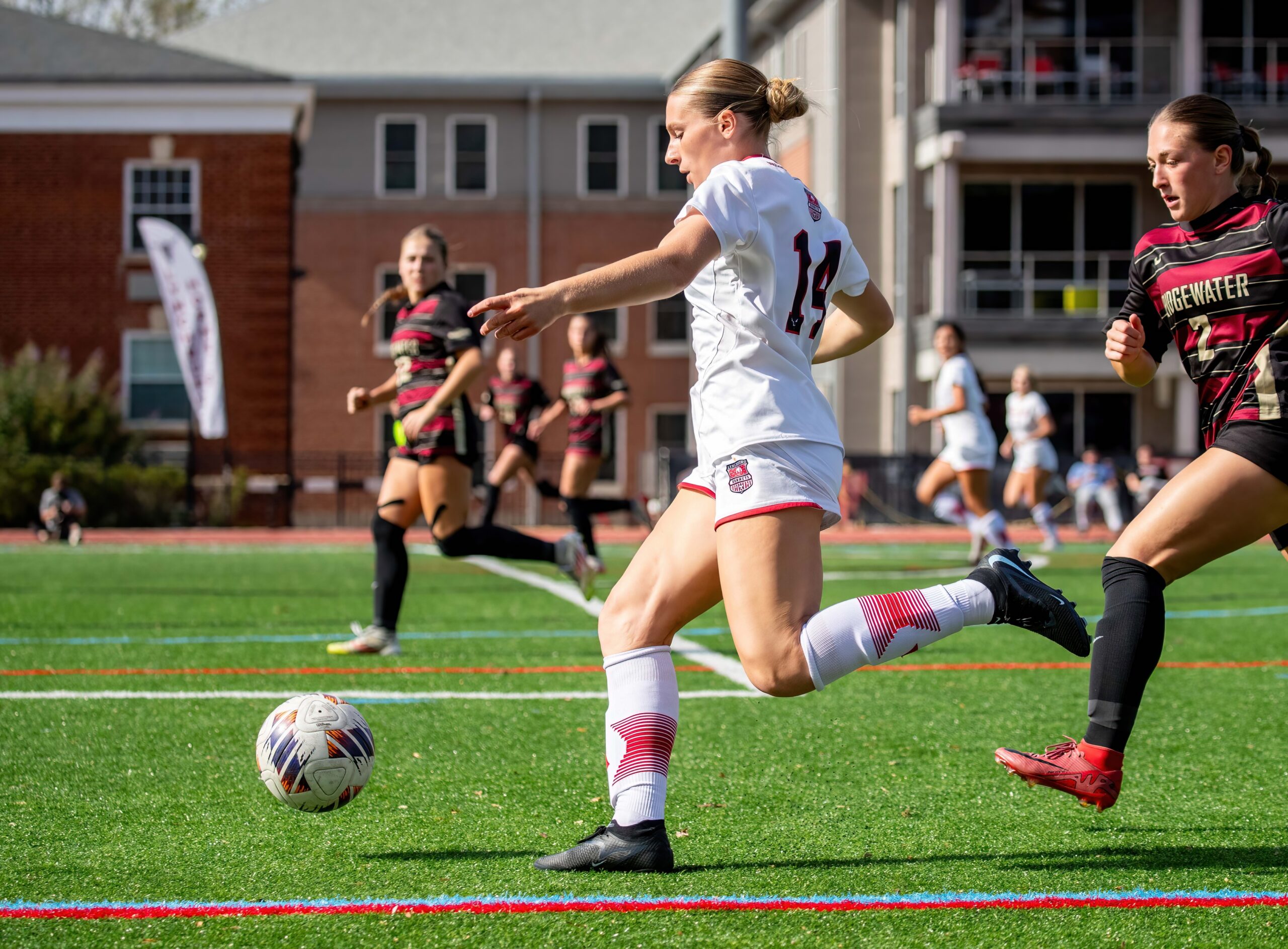 lynchburg women's soccer player in a white jersey chases after the ball and passes past an opposing defender.
