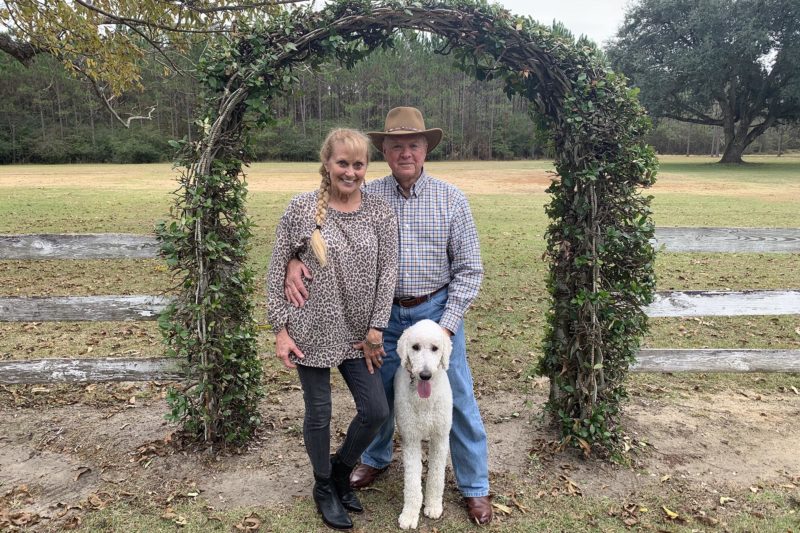 A middle-aged blond white woman and an elderly white man with cowboy hat and a white mid-sized dog stand in front of a worn white fence with farmland behind them
