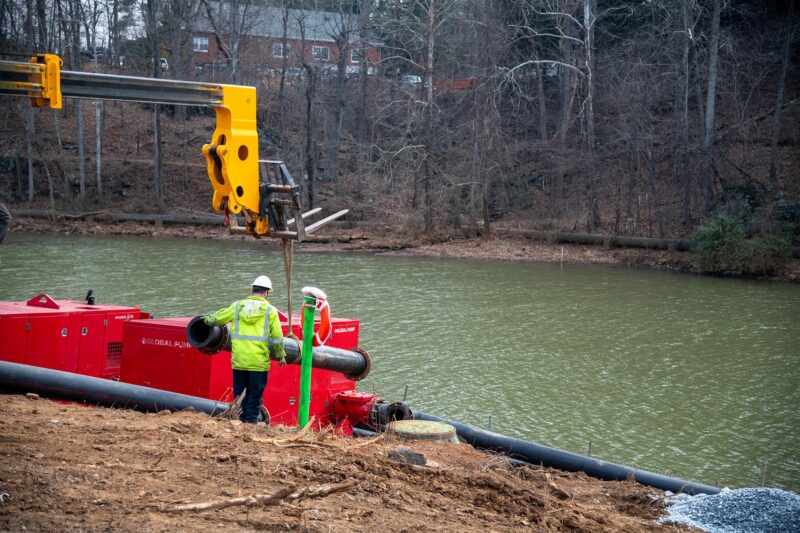 A construction worker holds a heavy pipe in front of a lake underneath a yellow and black forklift. A big red square metal pump can be seen in the background.