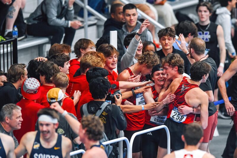 Young men in red track and field bibs hug and cry surrounded by fans